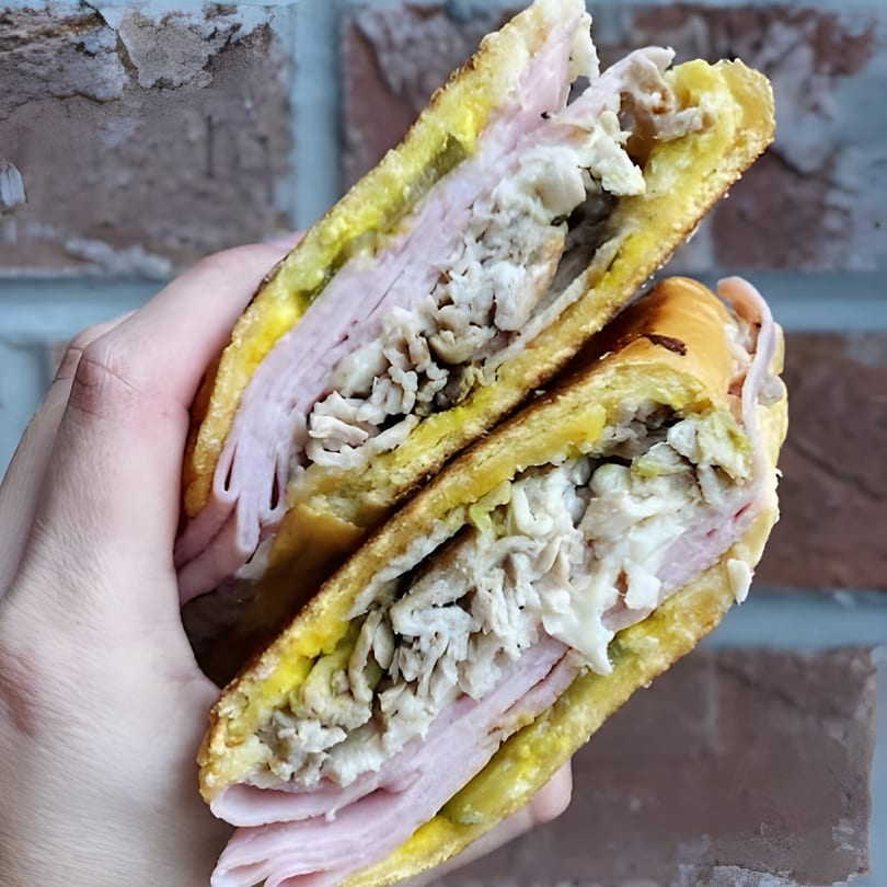A hand holds a sliced Cubano sandwich, revealing layers of ham, roasted pork, and pickles.