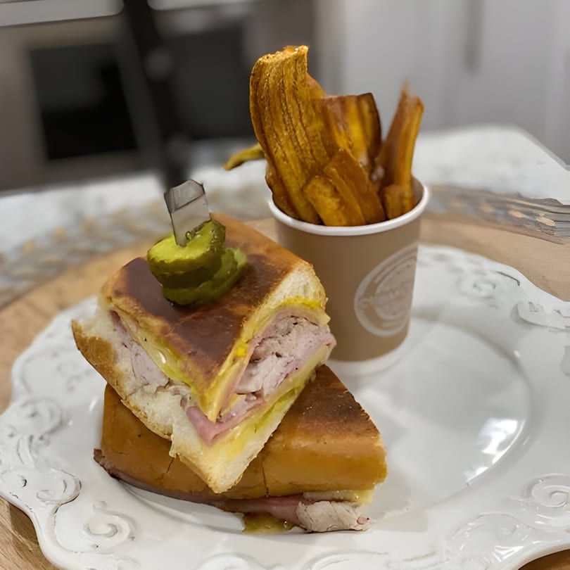 A Cubano sandwich with a pickle spear and a side of plantain fries in a branded cup.