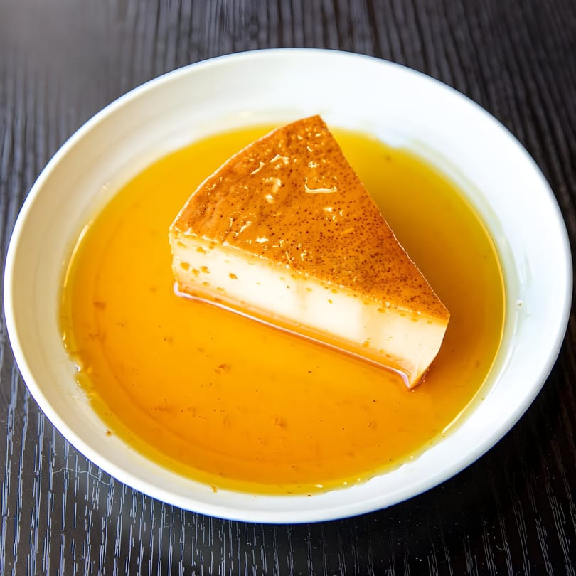 A slice of Cuban flan with caramel sauce in a white bowl on a dark wooden table.